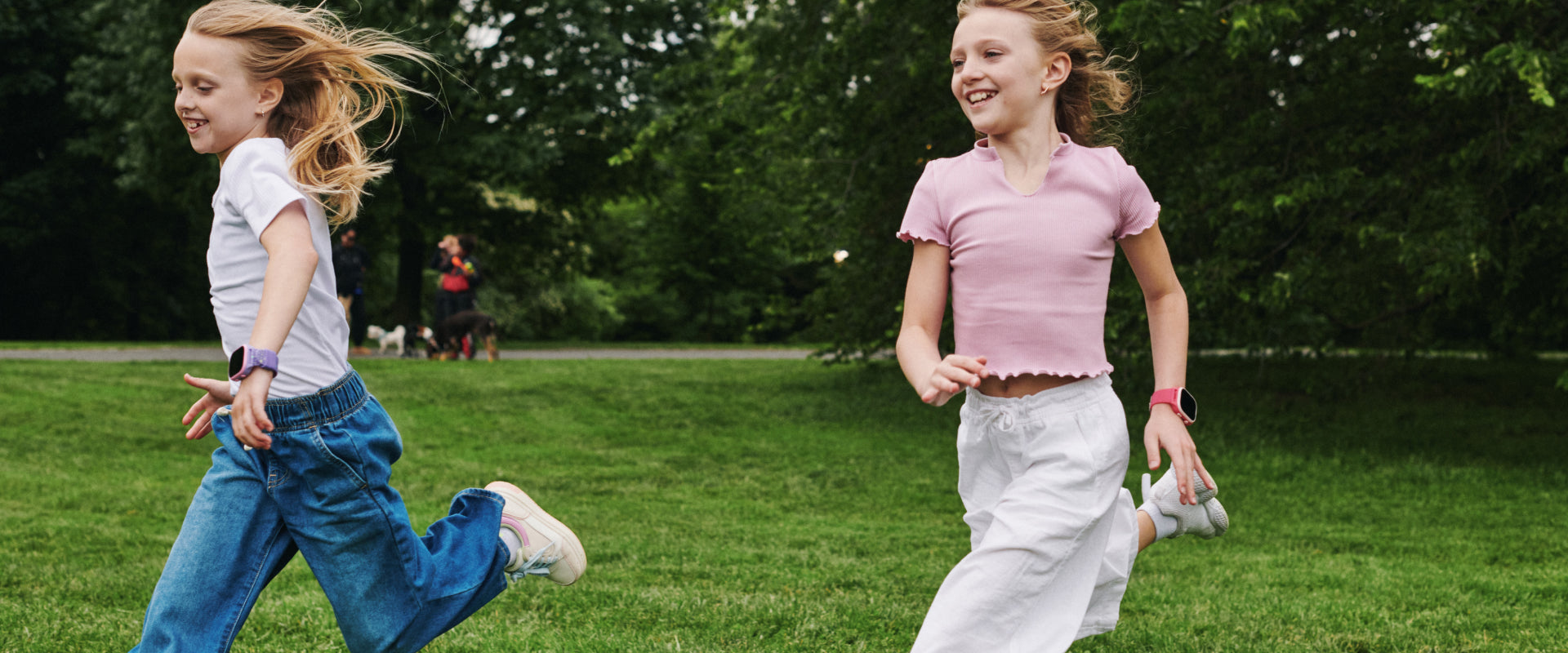 Two young girls running on a grassy field with trees in the background wearing smartwatches